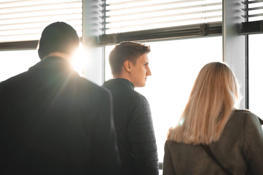 Personen stehen vor einem Fenster. Die Sonne scheint am Kopf der am weitesten links stehenden Person durch.