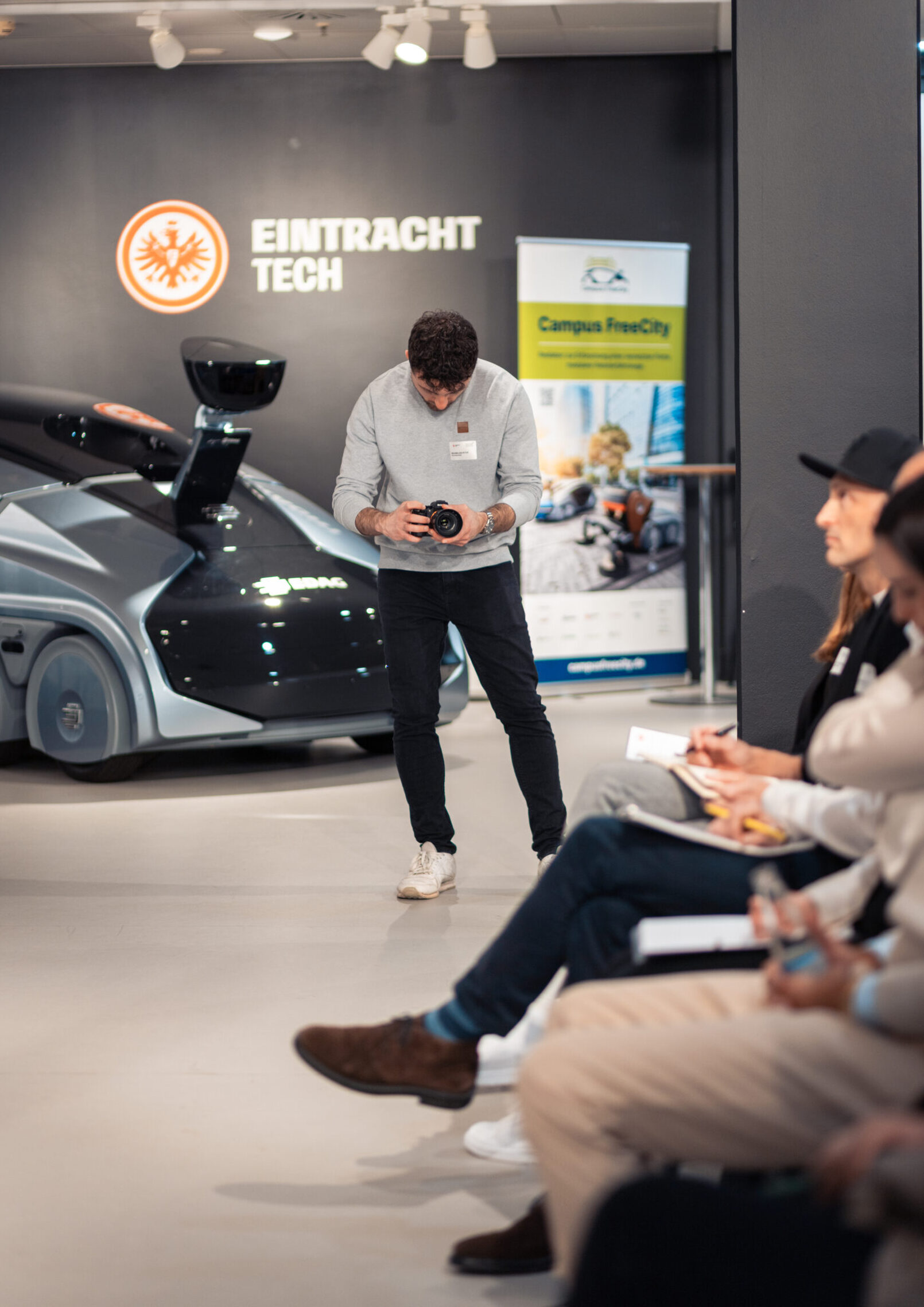 Fotograf während eines Events in Frankfurt am Main fotografiert. Er hat einen direkten blick in die Kamera, ist seitlich zu dieser gedreht, während das Event im Hintergrund stattfindet