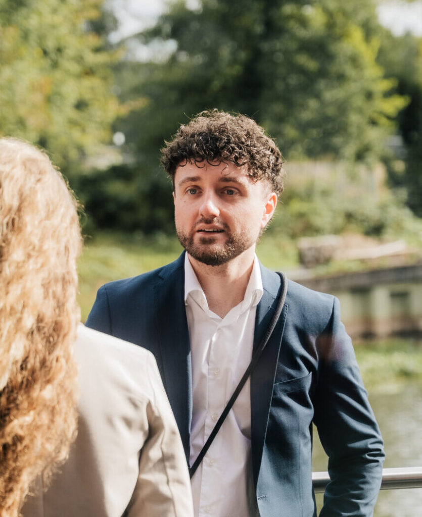 Das Foto zeigt den gutaussehenden Eventfotografen selbst bei einem Business-Event. Er unterhält sich mit anderen Teilnehmenden Personen auf dem Event. Man sieht seine Kamera im Vordergrund
